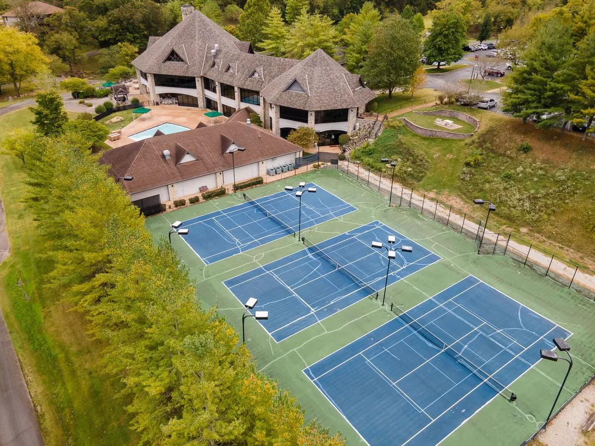 Modern tennis courts adjacent to a clubhouse, surrounded by neatly landscaped grounds and greenery. The setting is bright and energetic, hinting at ongoing tennis matches and social gatherings.