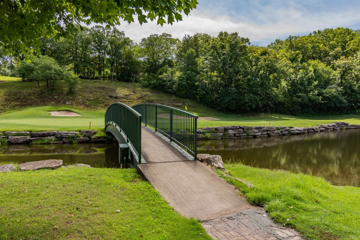 A scenic view of a green bridge crossing calm water, surrounded by a golf course and lush trees. The area appears tranquil and inviting—perfect for leisurely walks, nature observation, or photographing wildlife, with the landscape rolling gently into the distance.