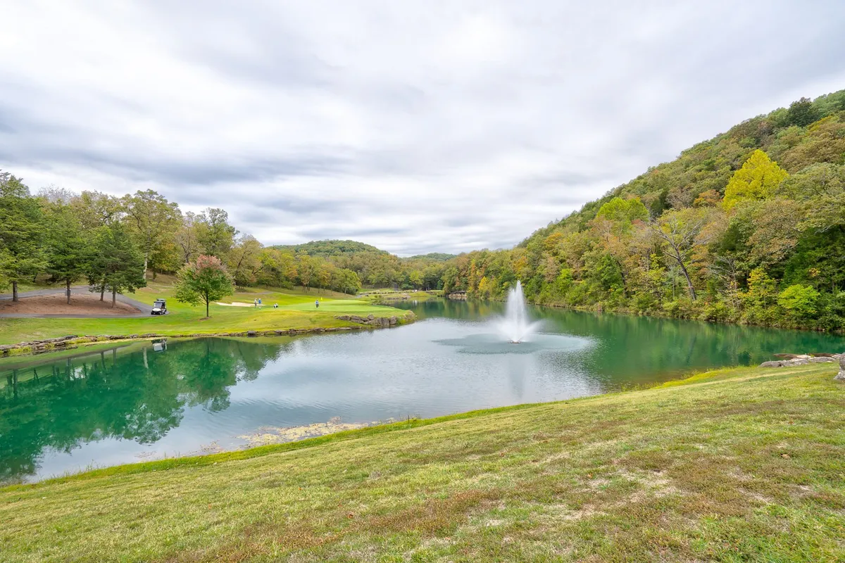 A picturesque view of a lake featuring a sparkling fountain, surrounded by rolling hills and abundant greenery. The peaceful setting highlights opportunities for fishing, walking, and nature observation, with the sunlight dancing on the water and wildlife often present nearby.