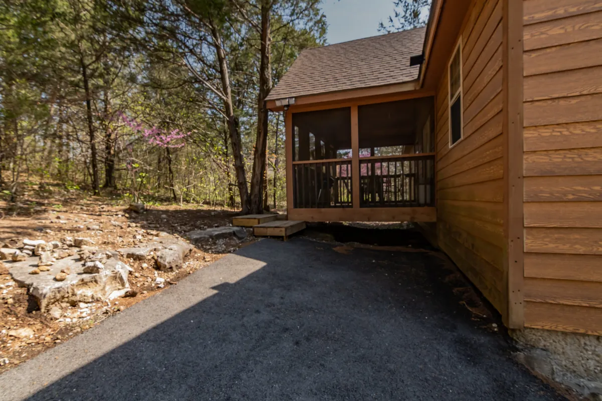 This image captures a screened porch nestled in natural surroundings, with a clear view of the porch structure, comfortable seating, and the greenery just beyond the screens. A walking path can be seen nearby, further integrating the porch with the outdoors. The lighting and ambiance suggest a tranquil, peaceful setting ideal for relaxation.