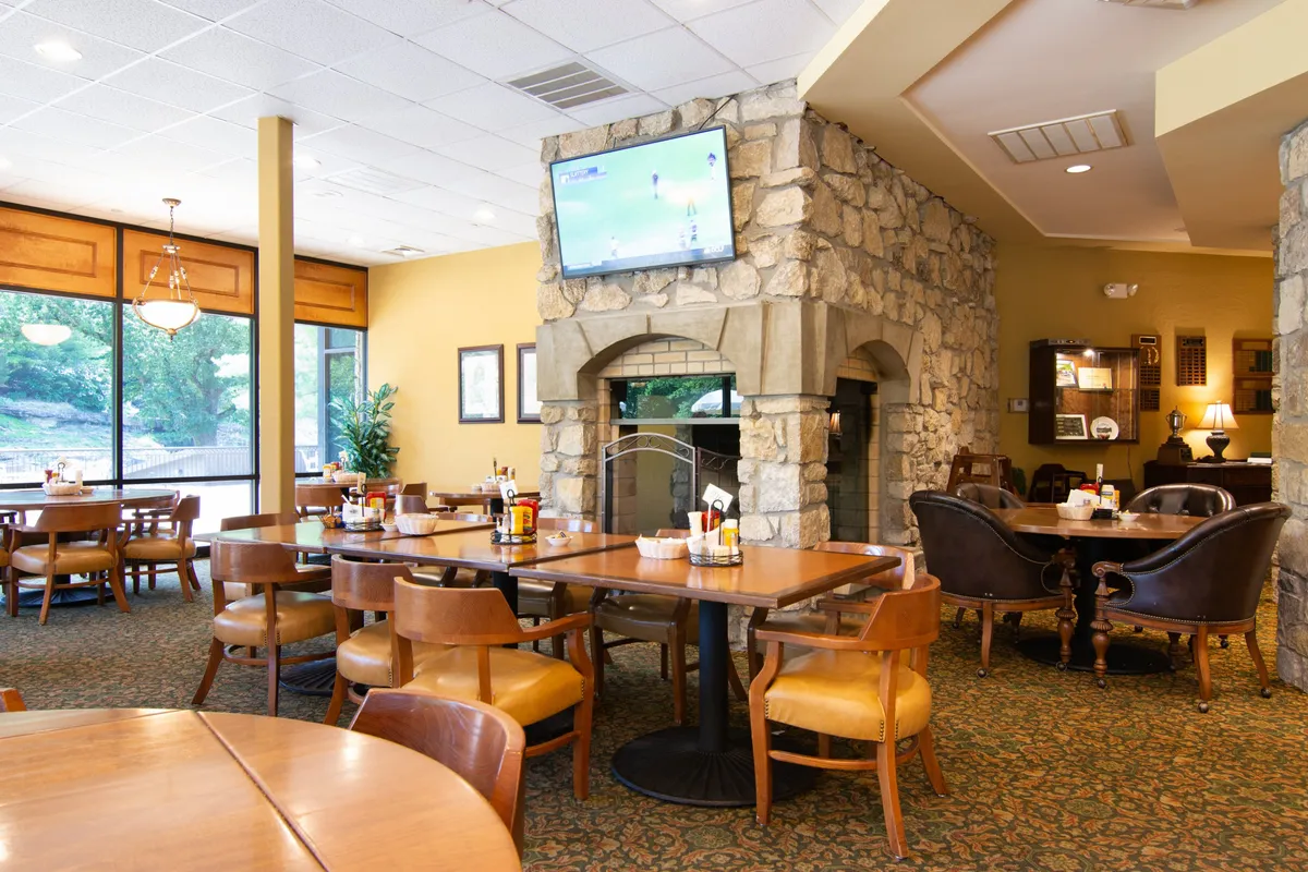 A cozy interior of the Ledgestone Grille, featuring a rustic stone fireplace, wooden tables, and large windows that flood the space with natural light. The TV screen and communal seating create a welcoming, social vibe ideal for post-golf relaxation.