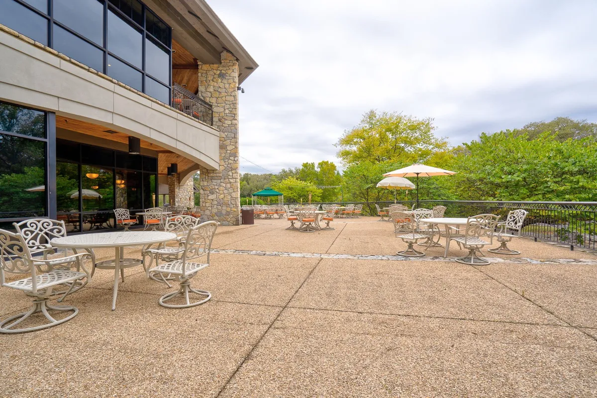 An elegant exterior scene showcasing an outdoor seating area adjacent to a stone building facade, framed by vibrant greenery. The area is set up for social gatherings or casual outdoor meals, and the gentle lighting hints at the warmth of sunset. Subtle seasonal decorations add a welcoming, celebratory touch.