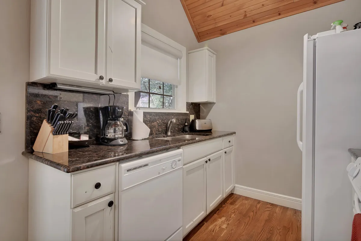 This image shows a warmly lit kitchen with granite countertops, modern appliances, and wooden cabinetry. The kitchen is spacious enough for several people to gather, and the natural light streaming through the window enhances its inviting and functional atmosphere.