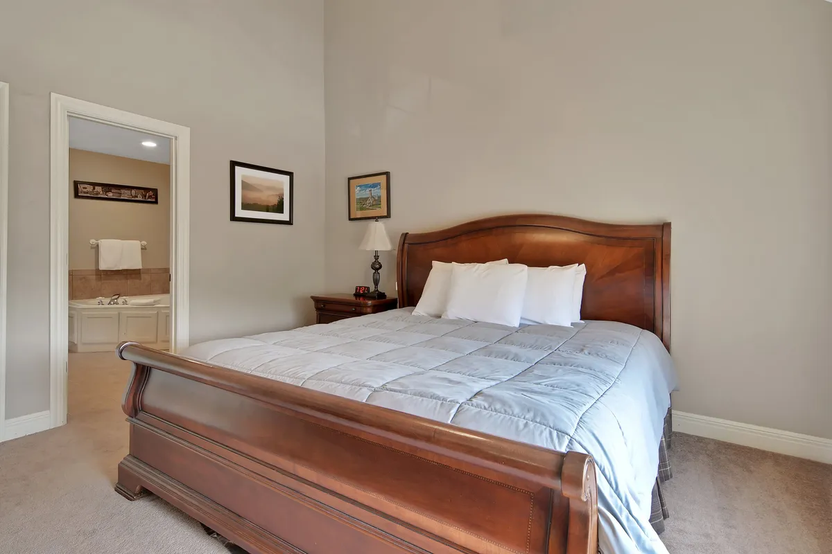 A tranquil bedroom featuring a wooden bed, decorative lamp, framed pictures, and a view into an adjoining bathroom. The calming ambiance and comfortable setup invite relaxation and restful nights.
