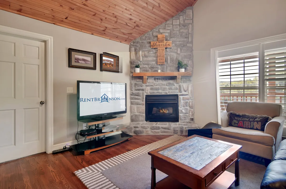 A cozy living room with a stone fireplace, a TV atop a wooden media stand, a soft couch with decorative cushions, and a wooden coffee table. The space is arranged for comfort, socializing, and quiet relaxation.