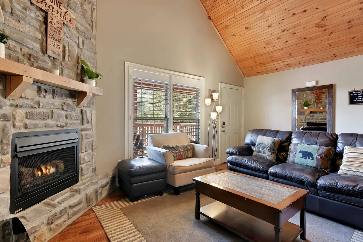 A welcoming living room featuring a striking stone fireplace, leather sofas arranged around a wooden coffee table, and large windows that let in natural light. The space exudes warmth and comfort, perfect for relaxing, socializing, or enjoying a cozy night in.