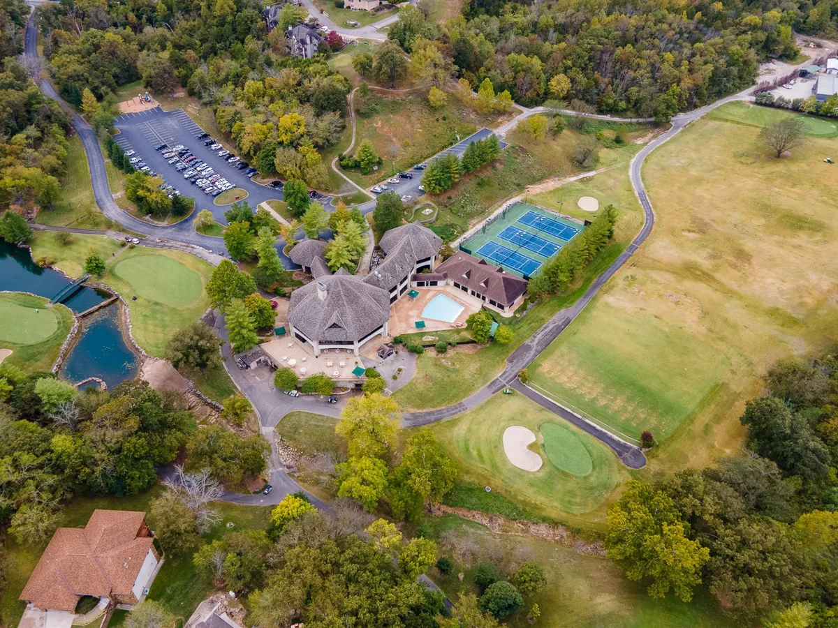 An expansive exterior view showing the main clubhouse, swimming pool, tennis courts, and a glimpse of the golf course, all nestled in verdant greenery. The gathering spaces suggest a vibrant resort atmosphere with several amenities visible.