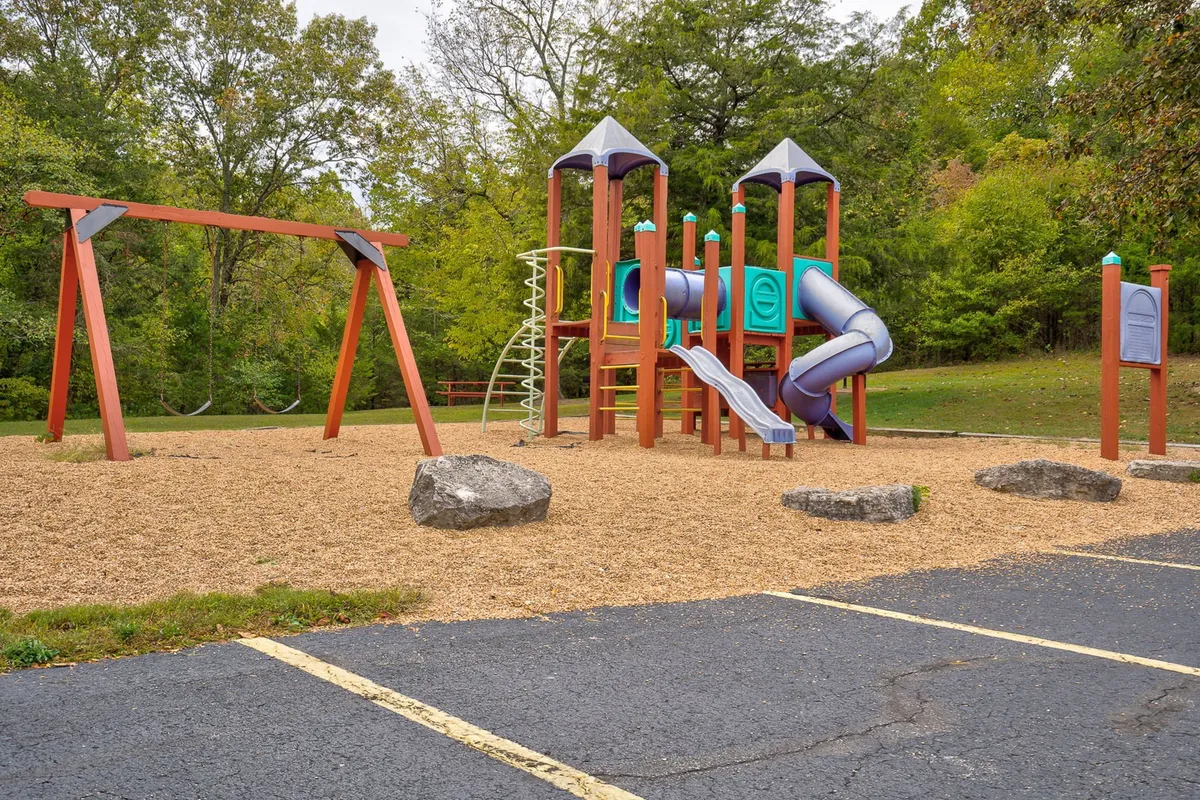 A vibrant playground structure with swings and climbing apparatus set in a grassy area surrounded by trees. Children can be seen playing, with families gathered nearby, creating a lively and joyful atmosphere.