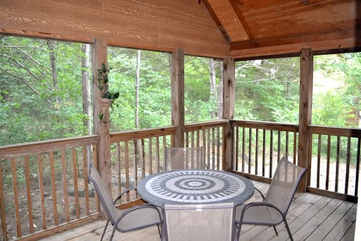 The image shows the interior of a screened-in porch with a wooden table featuring a patterned top, surrounded by trees visible through the screens and wooden railings. The natural light and textures highlight a cozy dining or gathering space that feels immersed in the surrounding woods, reinforcing the idea of nature's living room.